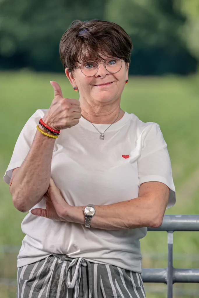 Eine lächelnde Frau mit Brille und kurzen dunklen Haaren steht im Freien und zeigt motiviert den Daumen nach oben.