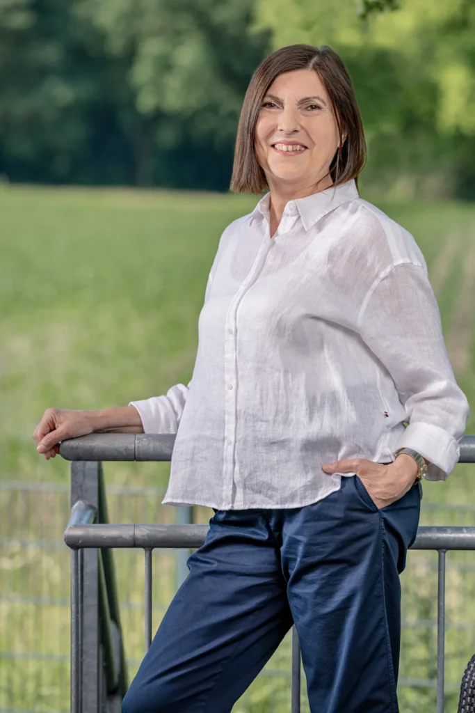 Eine sympathisch lächelnde Frau mit mittellangen braunen Haaren im weißen Leinenhemd steht entspannt an einem Metallgeländer im Freien.