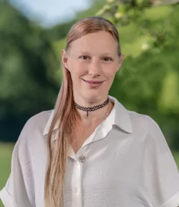 Eine freundliche junge Frau mit langen, rötlich-blonden Haaren und einer schwarzen Halskette steht in einer weißen Bluse lächelnd vor einer grünen Wiese.
