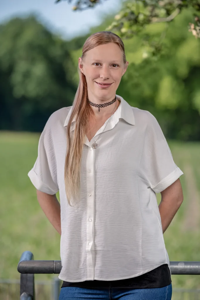 Eine freundliche junge Frau mit langen, rötlich-blonden Haaren und einer schwarzen Halskette steht in einer weißen Bluse lächelnd vor einer grünen Wiese.