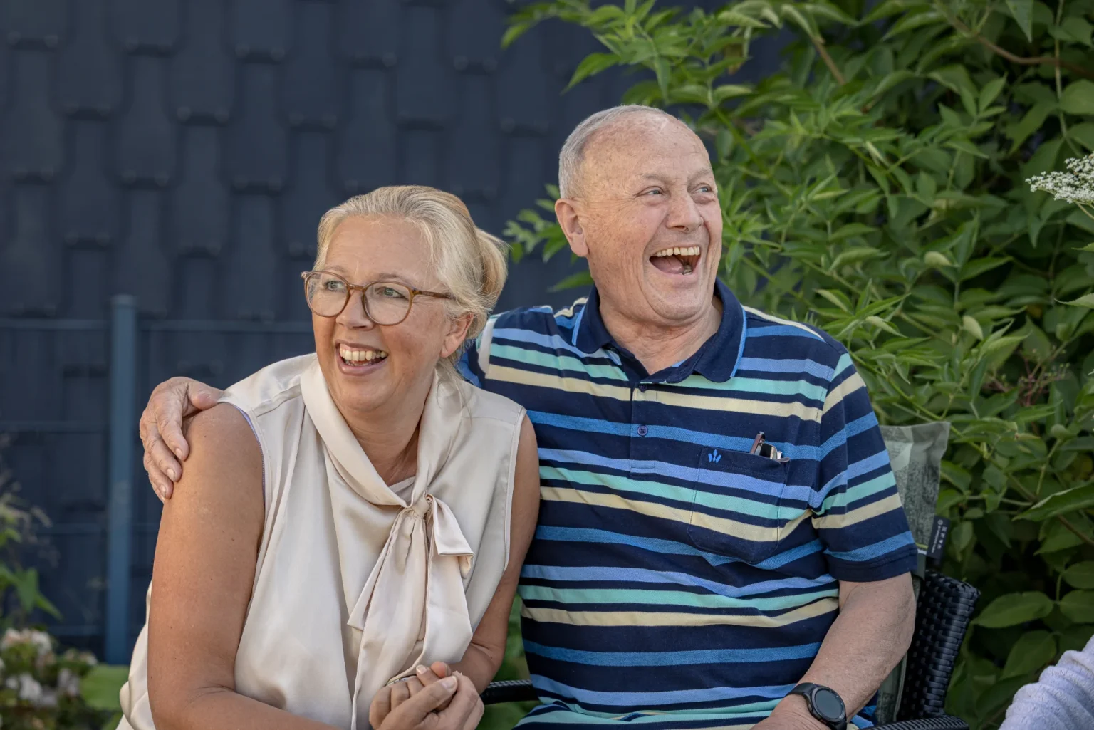 Eine lächelnde Frau mit Brille und ein herzlich lachender älterer Mann im gestreiften Poloshirt sitzen im Freien eng beieinander und halten sich an den Händen.