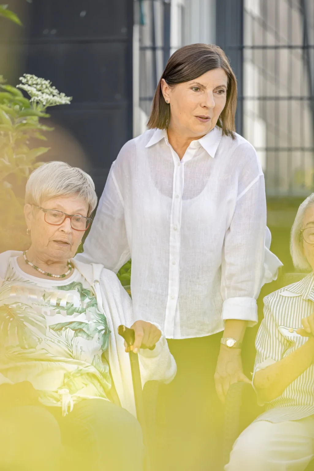 Pflegekraft im Gespräch mit zwei Seniorinnen im Garten der Tagespflege