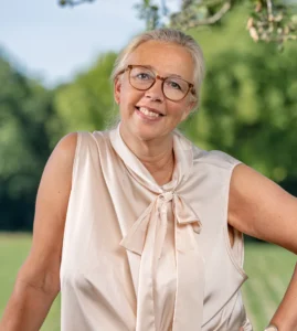 Eine freundlich lächelnde Frau mit Brille und hochgestecktem blonden Haar steht in einer eleganten, beigefarbenen Seidenbluse im Freien und stützt sich an einem Geländer ab.
